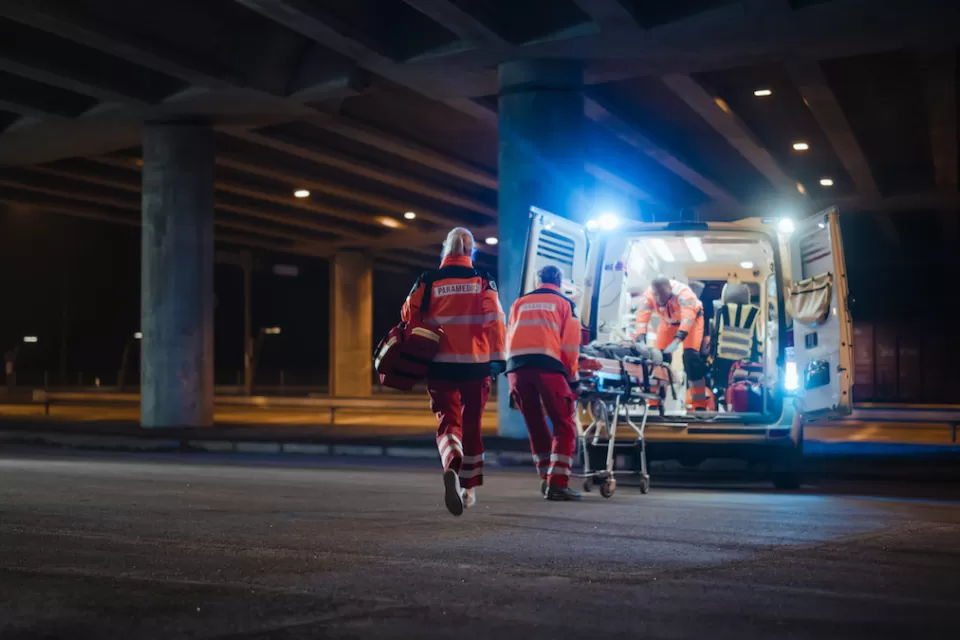 Three EMTs in orange uniforms putting gurney with person onto ambulance at night under freeway overpass.